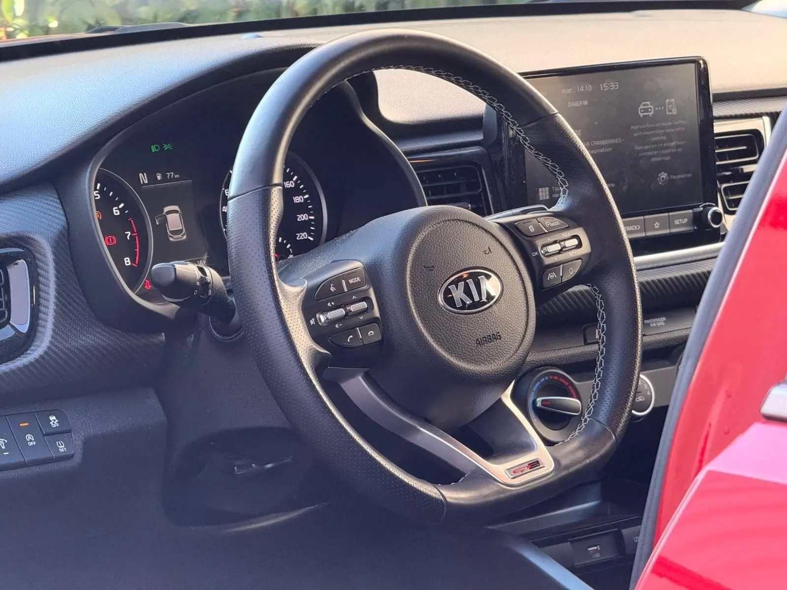 Vue intérieure centrée sur le volant cuir noir de la Kia Rio rouge grenadine, avec tableau de bord et écran tactile visibles.