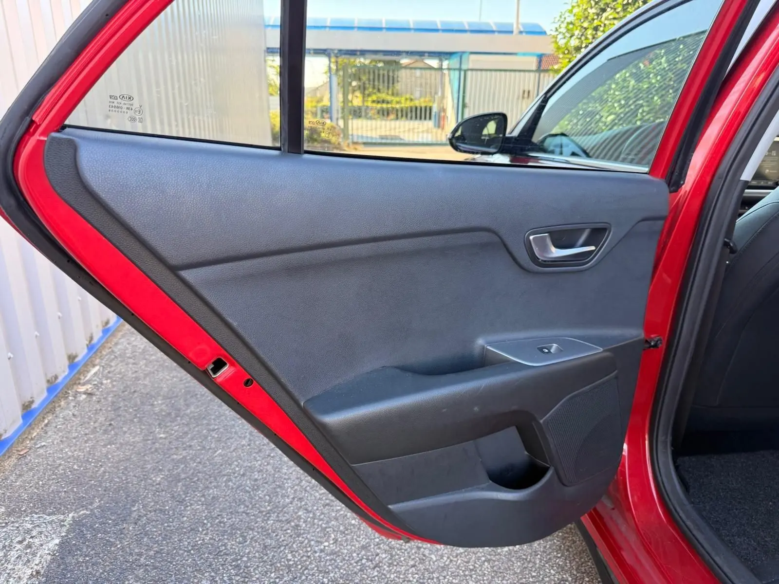 Porte arrière côté gauche ouverte d'une KIA RIO rouge grenadine 2022, intérieur noir avec commande de vitre électrique.