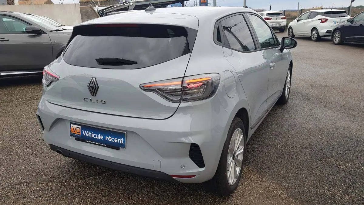 Vue 3/4 arrière droite d'une Renault Clio gris clair 2025 avec vitres surteintées et feux arrière allumés.