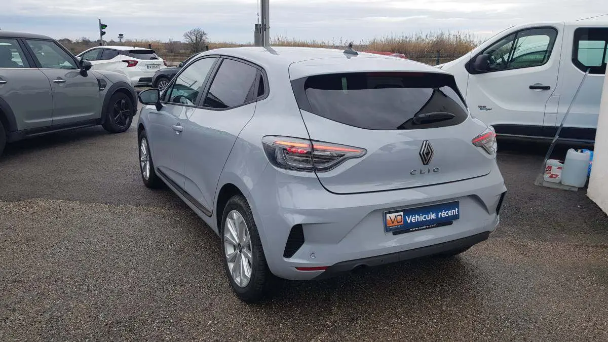 Vue 3/4 arrière d'une Renault Clio gris clair 2025 avec vitres surteintées et feux arrière allumés.