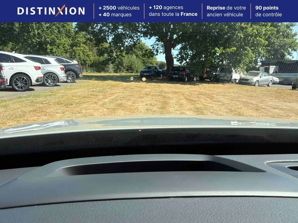 Vue depuis l'intérieur du Volvo XC90 gris 2024, montrant le tableau de bord et le pare-brise vers un parking extérieur.