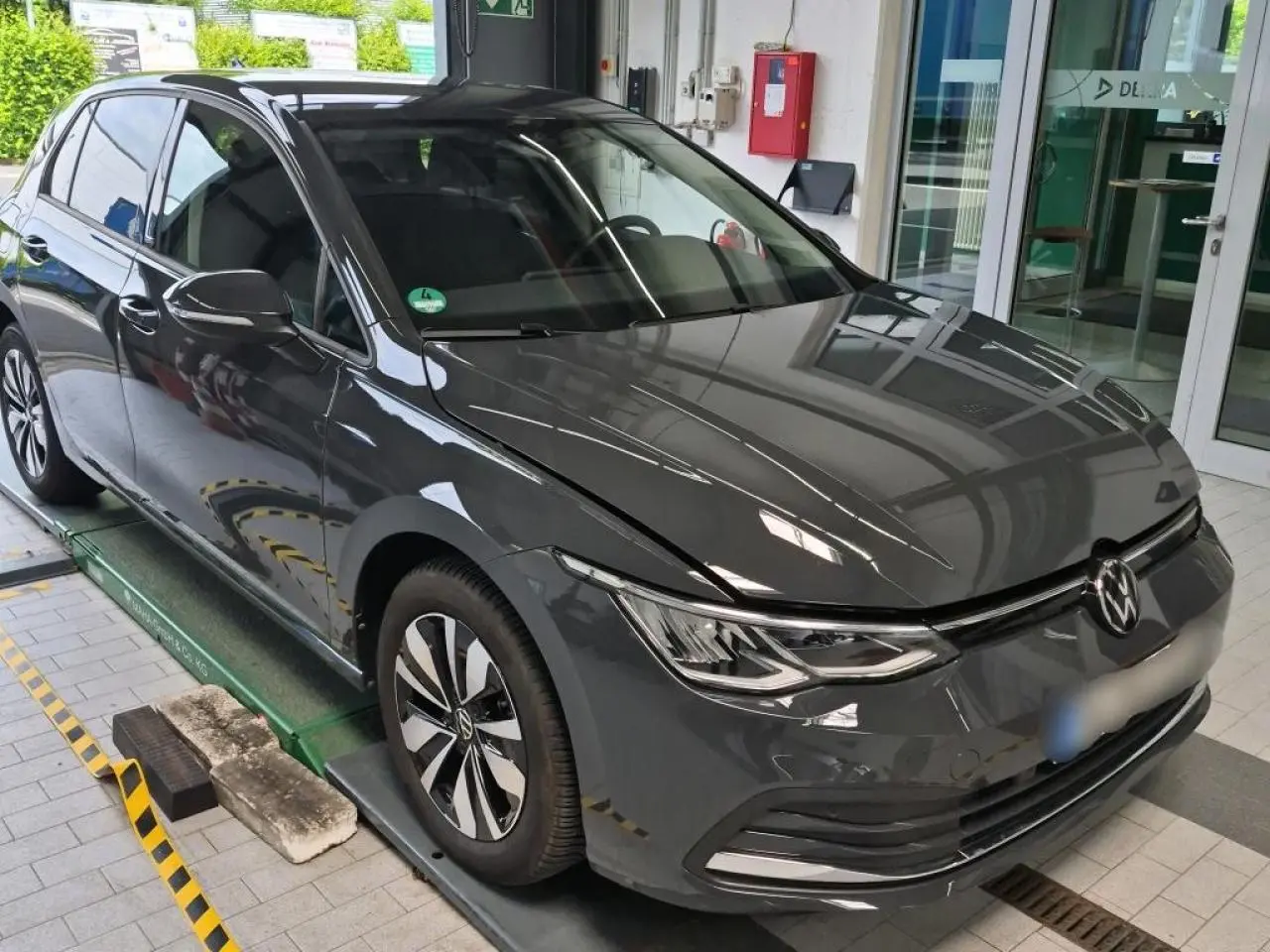 Volkswagen Golf 8 gris Urano vue 3/4 avant droit, avec jantes alliage et phares LED allumés en intérieur showroom.