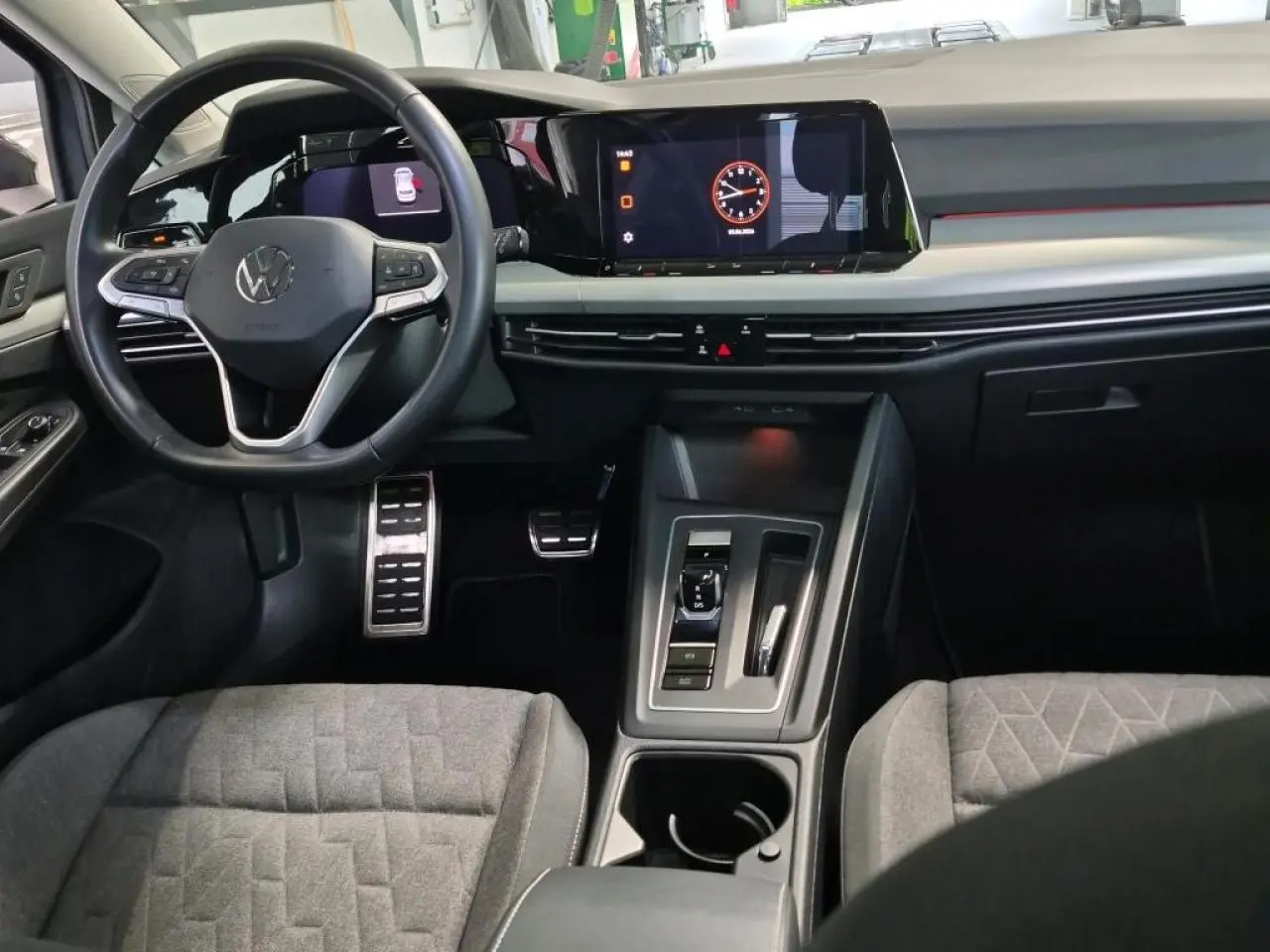 Vue intérieure avant de la Volkswagen Golf 8 avec tableau de bord numérique, volant multifonction et sellerie tissu gris.