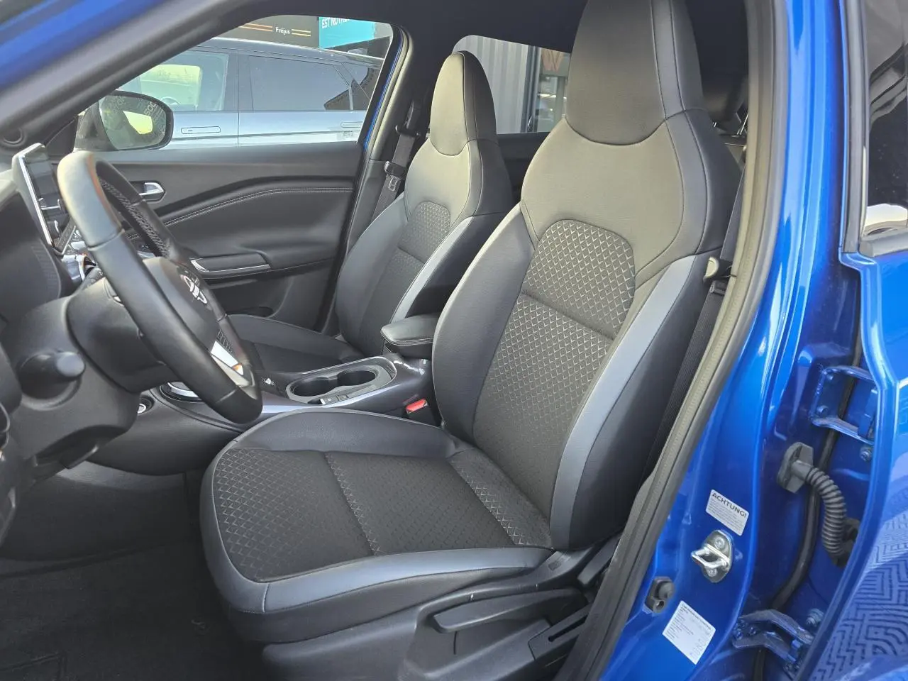 Vue intérieure côté conducteur du Nissan Juke II bleu magnetic, montrant les sièges avant en TEP noir et gris avec accoudoir central.