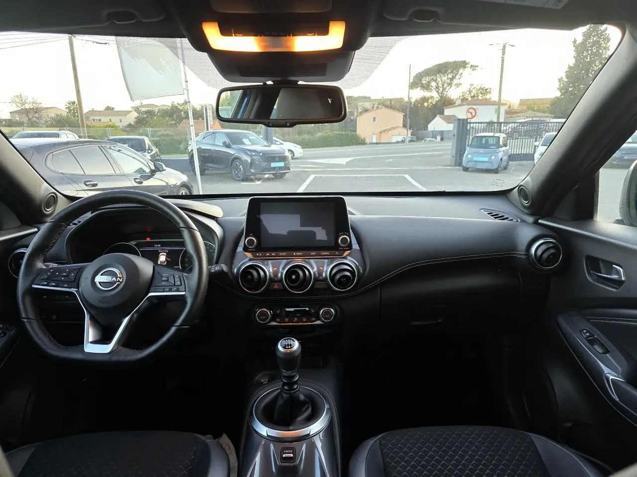 Intérieur noir du Nissan Juke II 2024 vu de face, avec volant cuir, écran tactile central et levier de vitesses manuel.