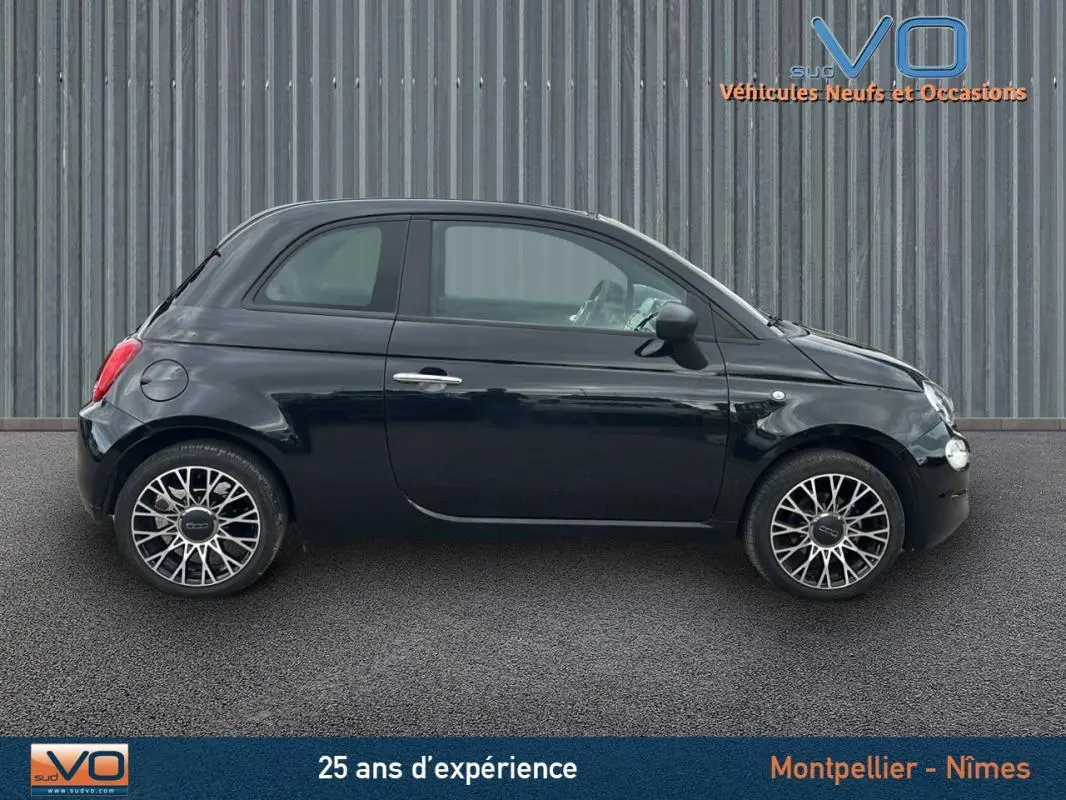 Profil côté gauche de la FIAT 500C noire 2024 avec roues alliage et roue de secours visible sur fond urbain.