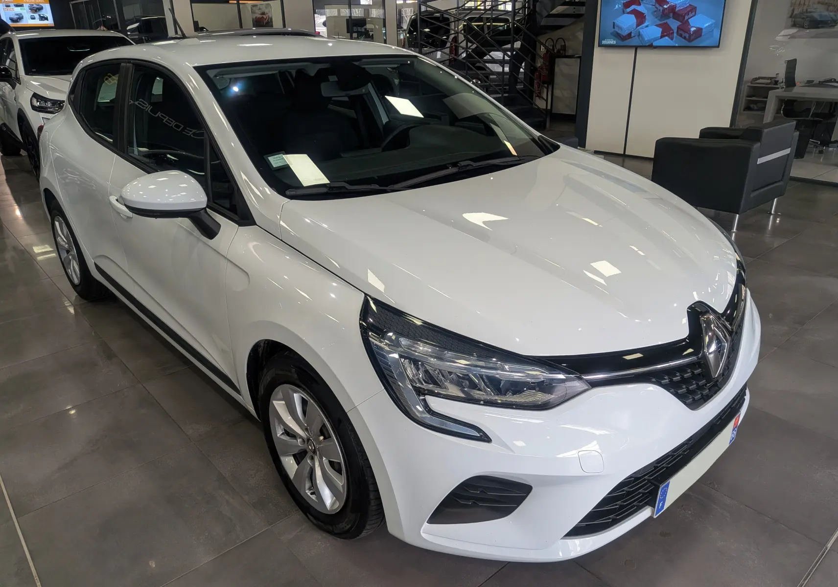 Vue 3/4 avant droite d'une Renault Clio V blanche Glacier en showroom, avec calandre noire et phares LED distinctifs.