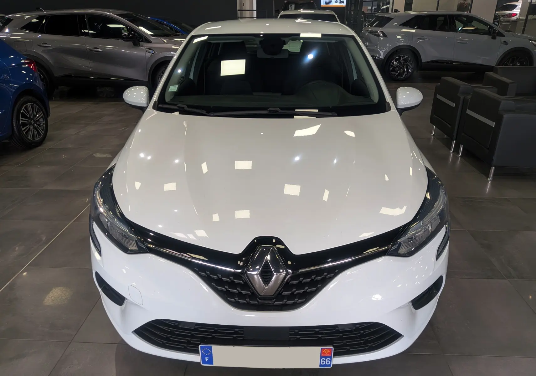 Vue avant d'une Renault Clio V blanche Glacier en showroom, mettant en valeur la calandre noire et le logo Renault central.