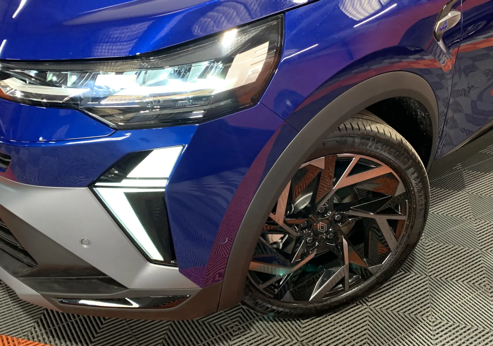 Gros plan sur l'avant droit bleu Iron du Renault Symbioz E-Tech, mettant en valeur le phare LED et la jante noire stylisée.