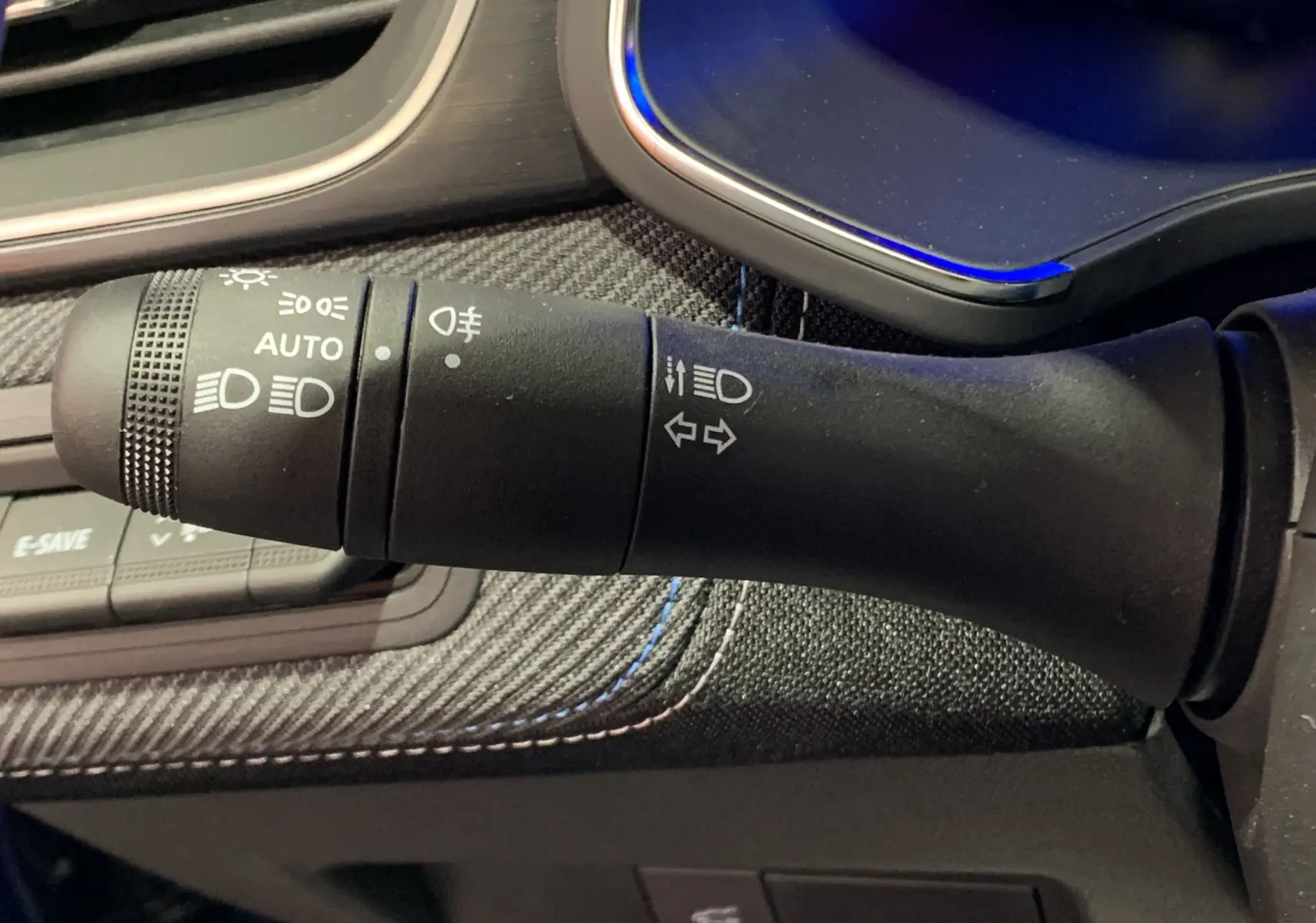 Gros plan sur la manette de commande des phares et clignotants, avec finition tissu noir et surpiqûres bleues du tableau de bord.