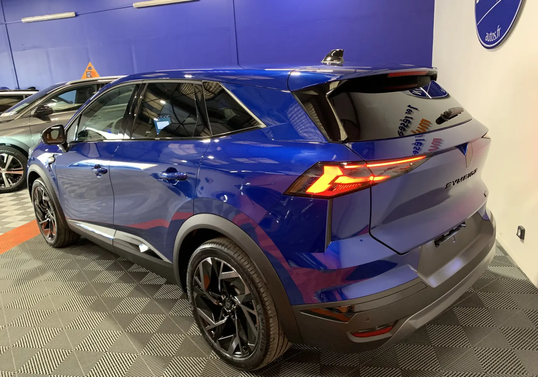 Vue 3/4 arrière droite d'un Renault Symbioz bleu Iron avec feux arrière allumés et jantes noires brillantes.