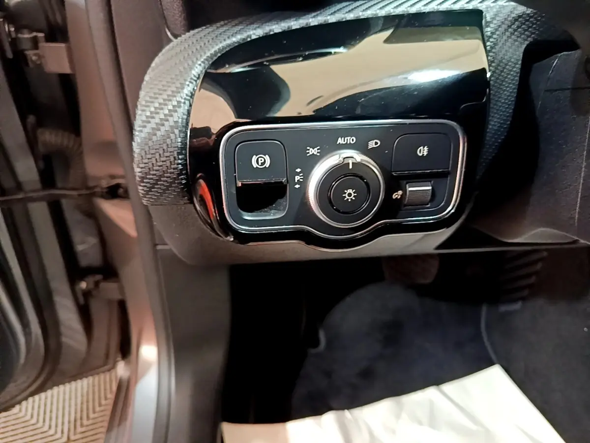 Gros plan sur le panneau de commande des phares et frein à main électrique côté gauche dans une Mercedes Classe A gris foncé.