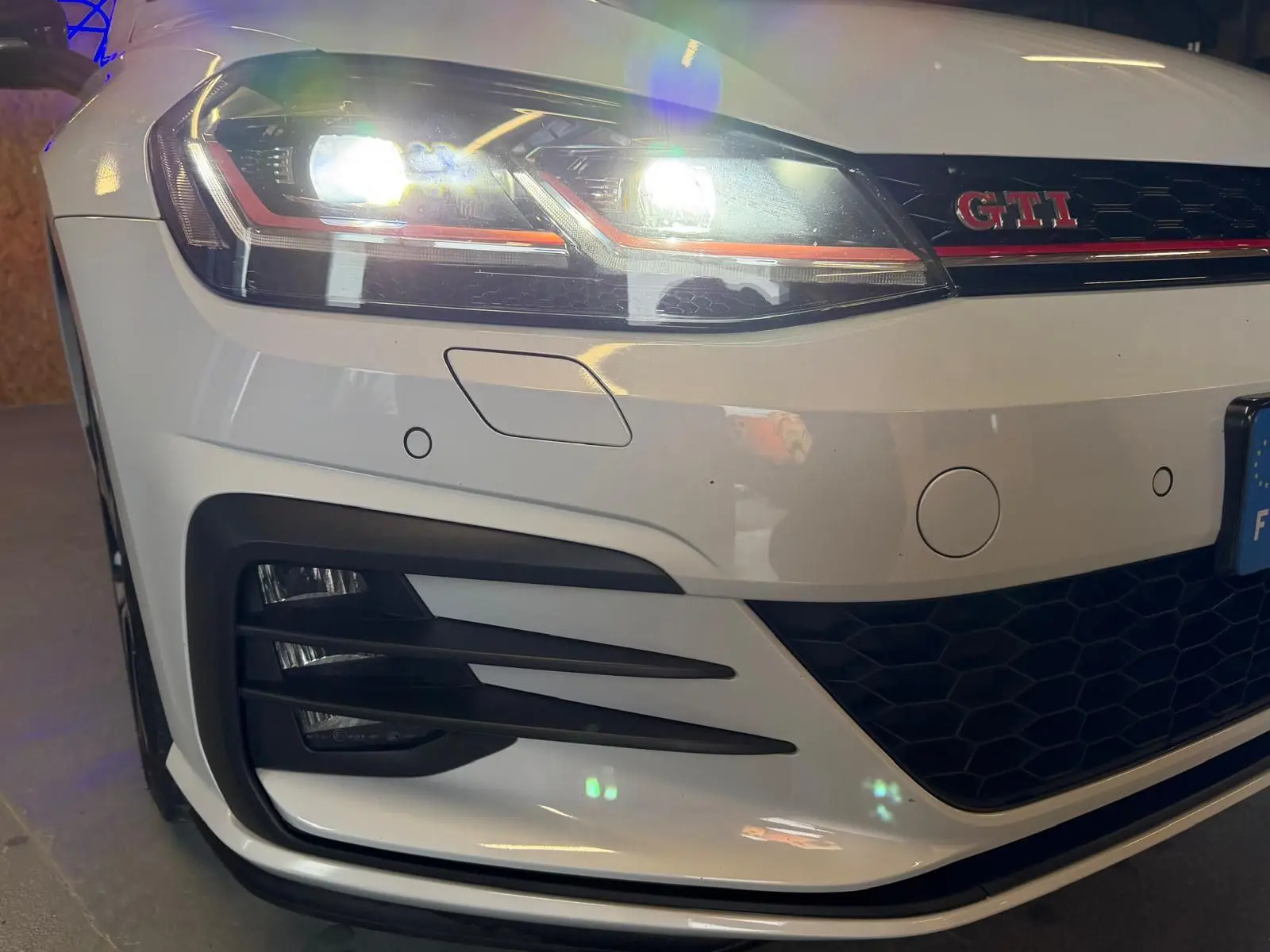 Gros plan avant gauche d'une Volkswagen Golf GTI Performance gris clair avec phares LED allumés et logo GTI rouge.
