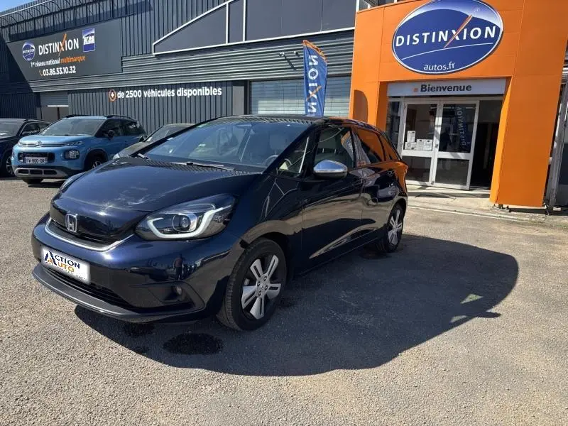 Honda Jazz 1.5 i-MMD bleu nuit nacré vue 3/4 avant droit, avec phares LED et jantes alu devant un garage Distinxion.