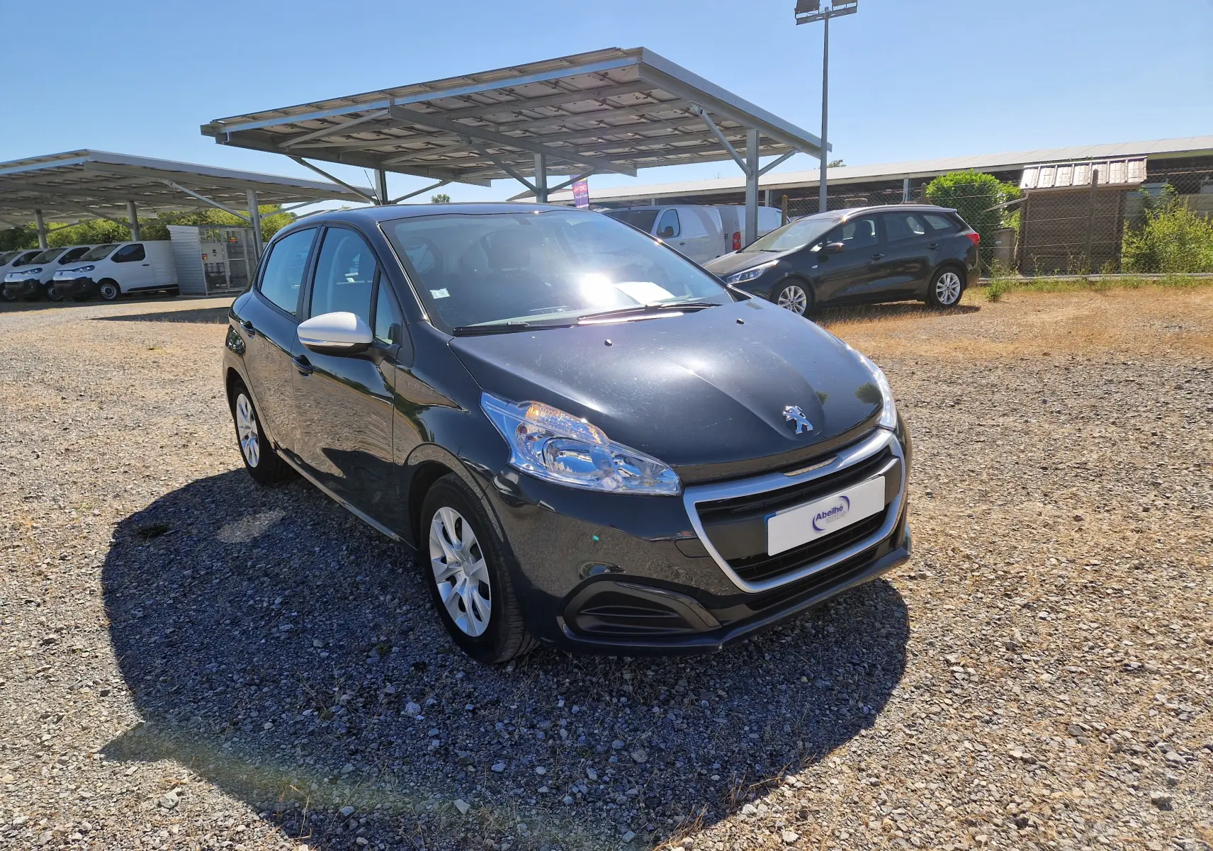 Peugeot 208 gris hurricane vue 3/4 avant droit sur parking, avec rétroviseurs blancs et jantes alliage.