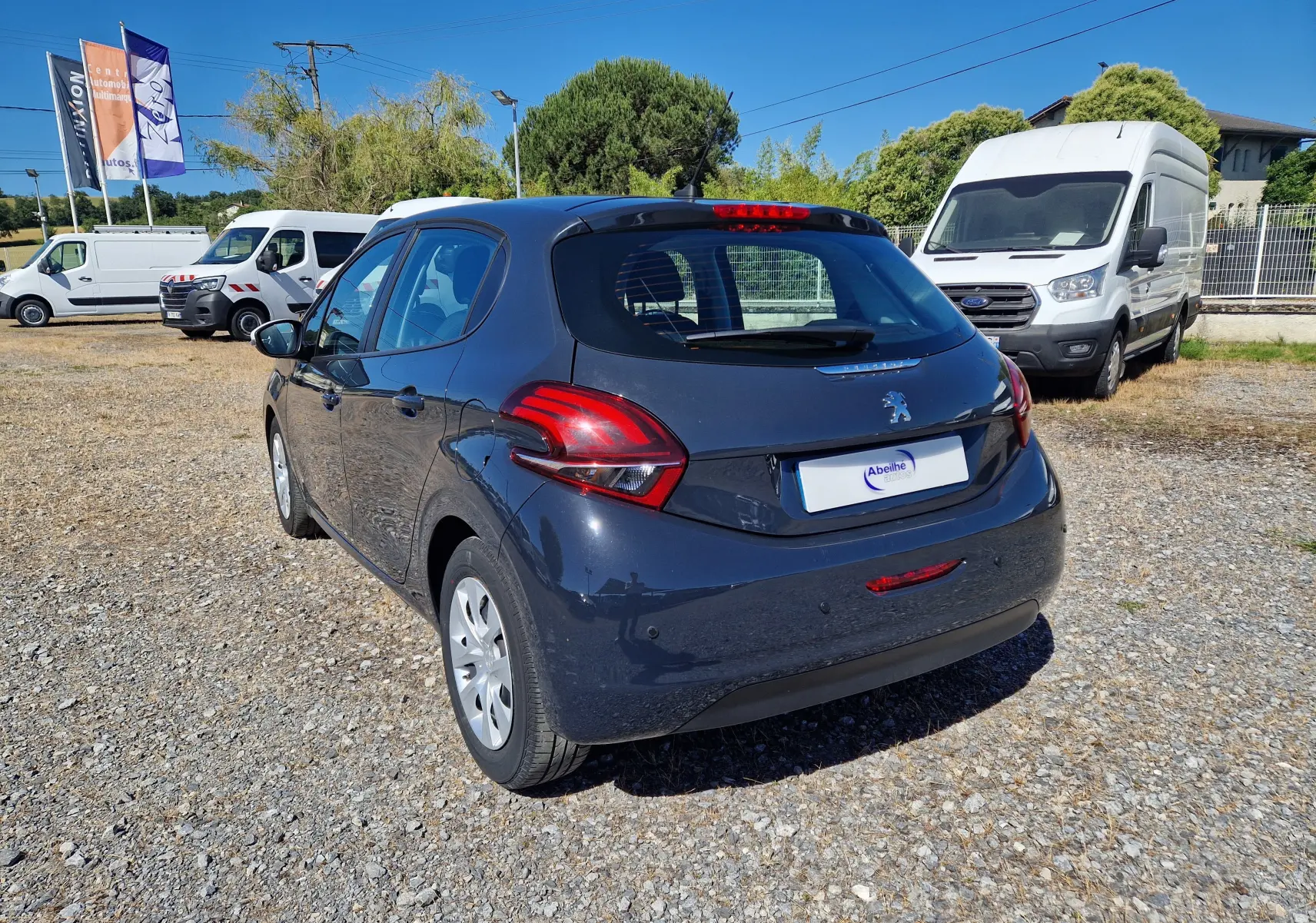Vue 3/4 arrière droite d'une Peugeot 208 gris hurricane 2018 garée sur un sol gravillonné, avec plusieurs véhicules utilitaires blancs en arrière-plan.