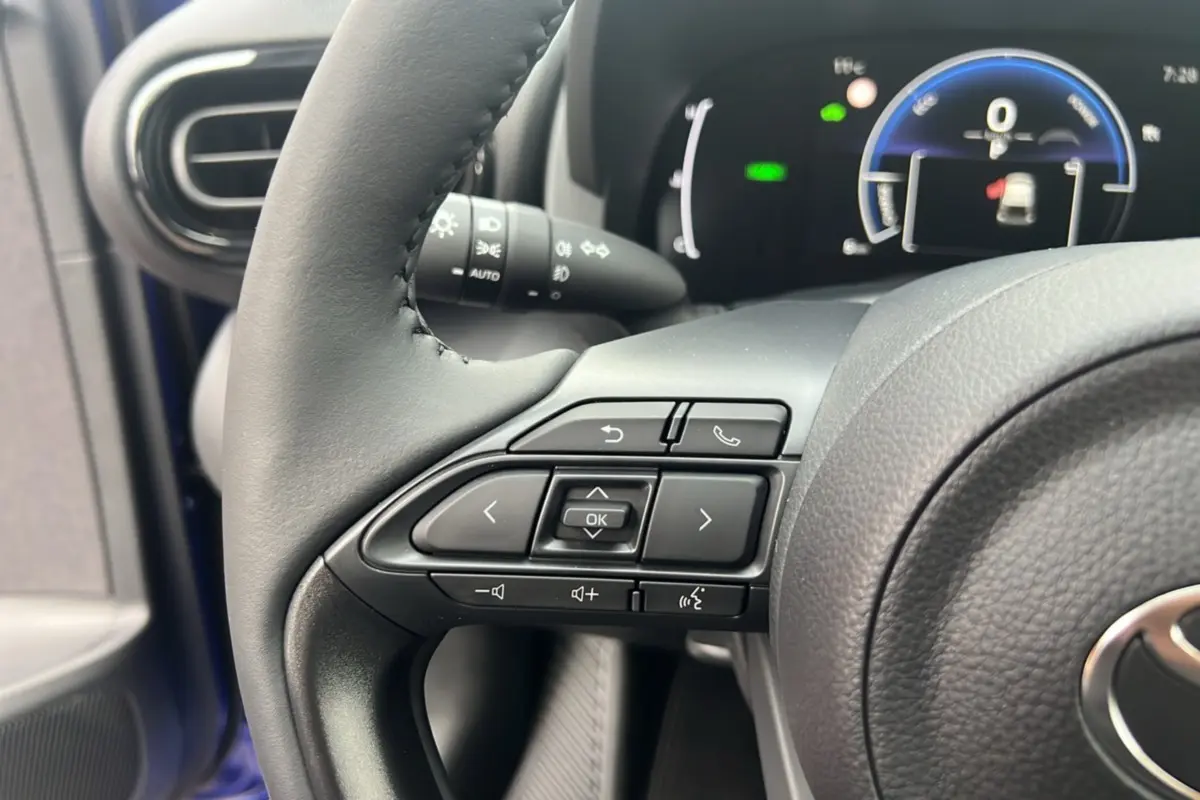 Gros plan sur les commandes au volant gauche de la Toyota Yaris 2025 bleu genievre, avec tableau de bord numérique en arrière-plan.