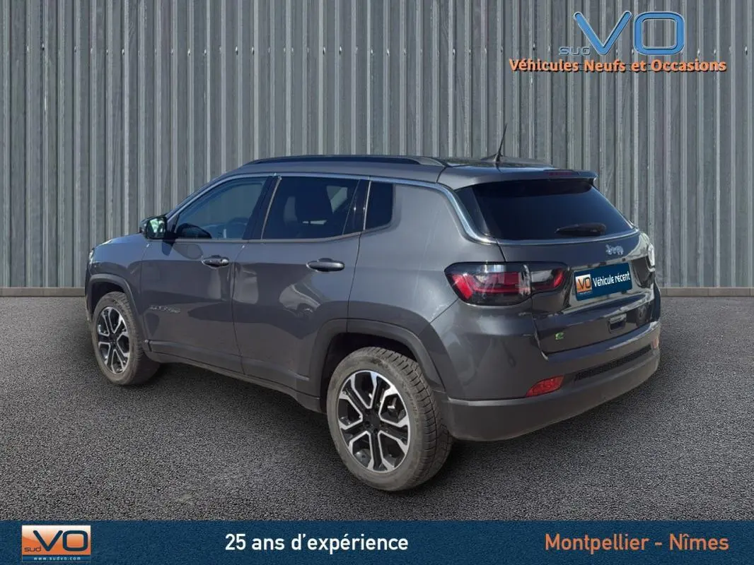 Jeep Compass gris 3/4 arrière droit avec barres de toit et jantes 18 pouces, stationné devant un mur métallique.