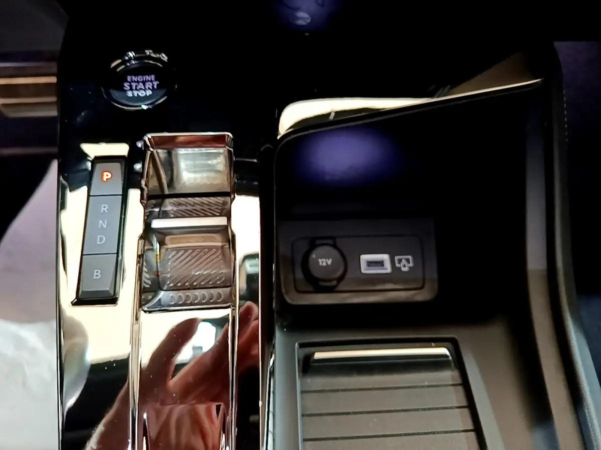 Gros plan sur la console centrale noire brillante de la Peugeot 308 PHEV 180 E-EAT8 GT avec levier de vitesses automatique et prise 12V.