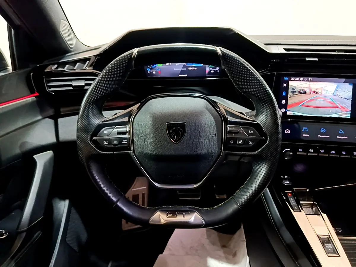 Vue intérieure centrée sur le volant cuir noir GT et le tableau de bord numérique de la Peugeot 308 PHEV 180 blanche.