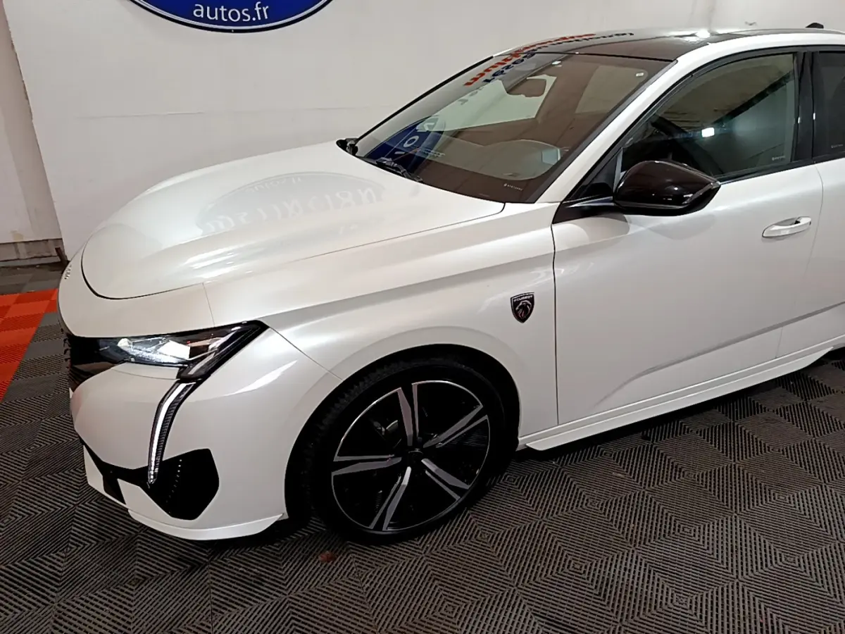 Vue 3/4 avant droite d'une Peugeot 308 PHEV 180 GT blanche avec jantes noires et logo GT sur aile avant.