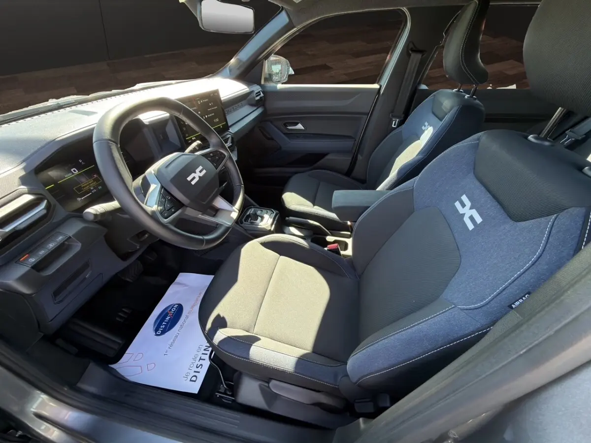 Vue intérieure côté conducteur du Dacia Duster gris foncé 2025, volant cuir multifonctions et sièges tissu bi-ton avec logo.