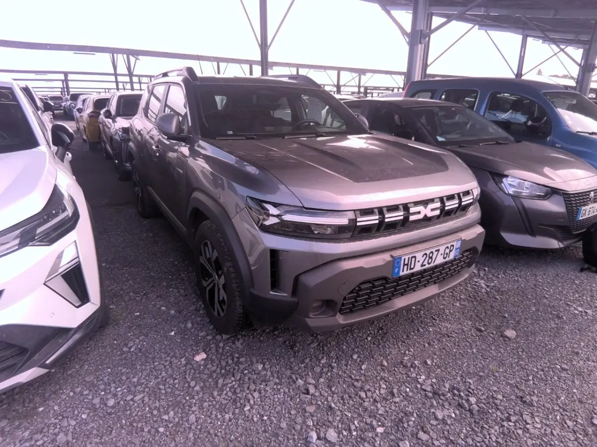Vue 3/4 avant d'un Dacia Duster gris foncé garé sous abri, avec calandre et phares LED distinctifs visibles.