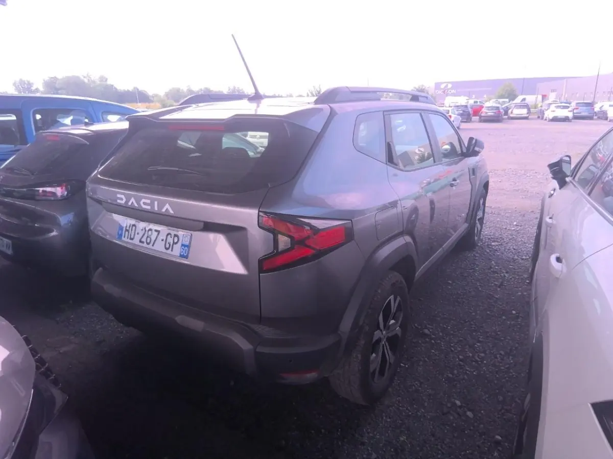 Vue 3/4 arrière droite d'un Dacia Duster gris foncé avec rails de toit noirs, stationné entre deux voitures.