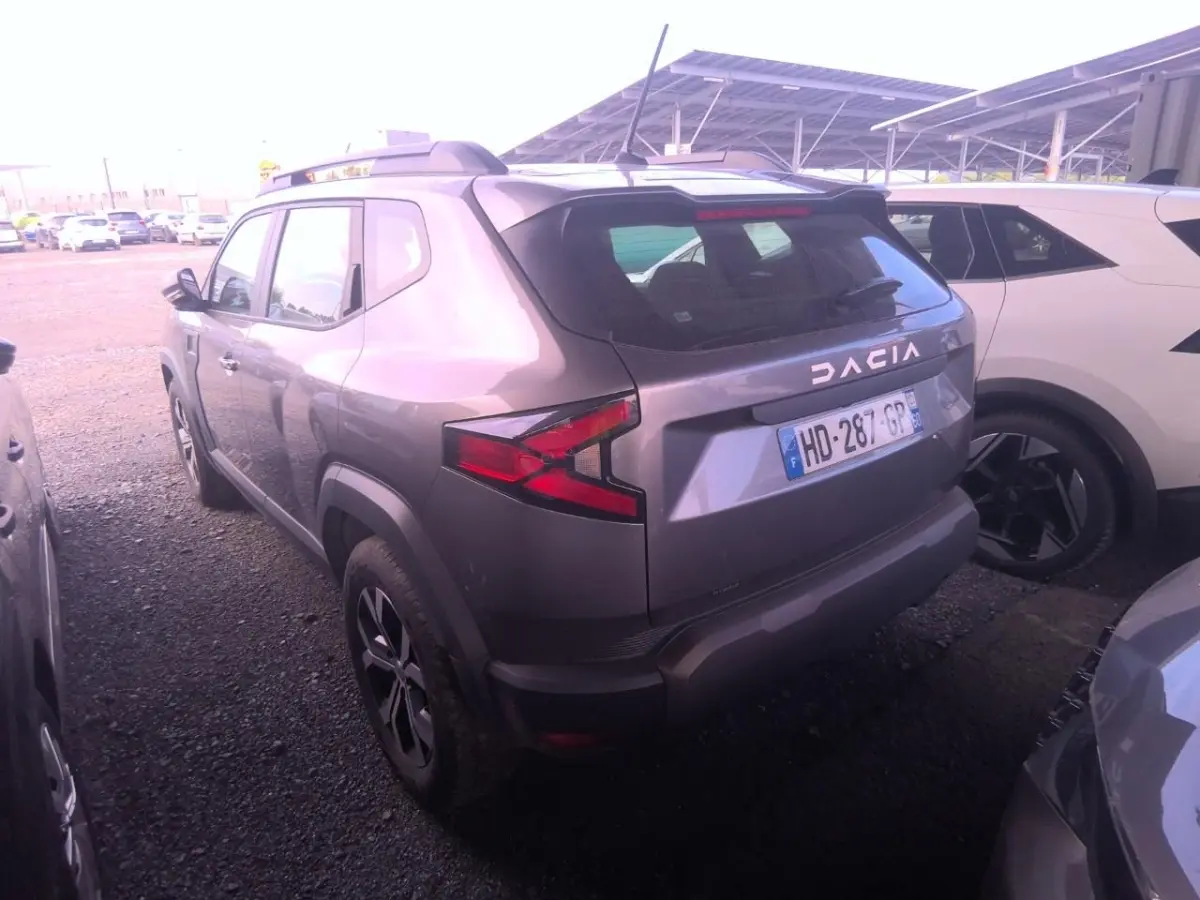 Vue 3/4 arrière d'un Dacia Duster gris foncé avec rails de toit noirs et feux arrière LED, stationné entre deux véhicules.