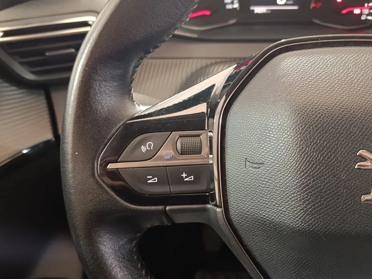 Gros plan sur le volant cuir noir de la Peugeot 208 blanc, avec commandes audio et bouton molette à gauche.