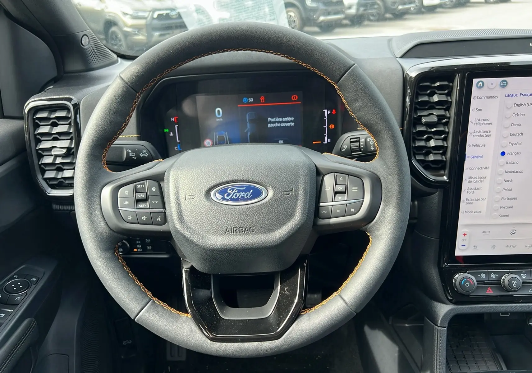 Vue centrée du volant en cuir noir avec surpiqûres orange du Ford Ranger 2025, tableau de bord numérique et écran tactile visible.