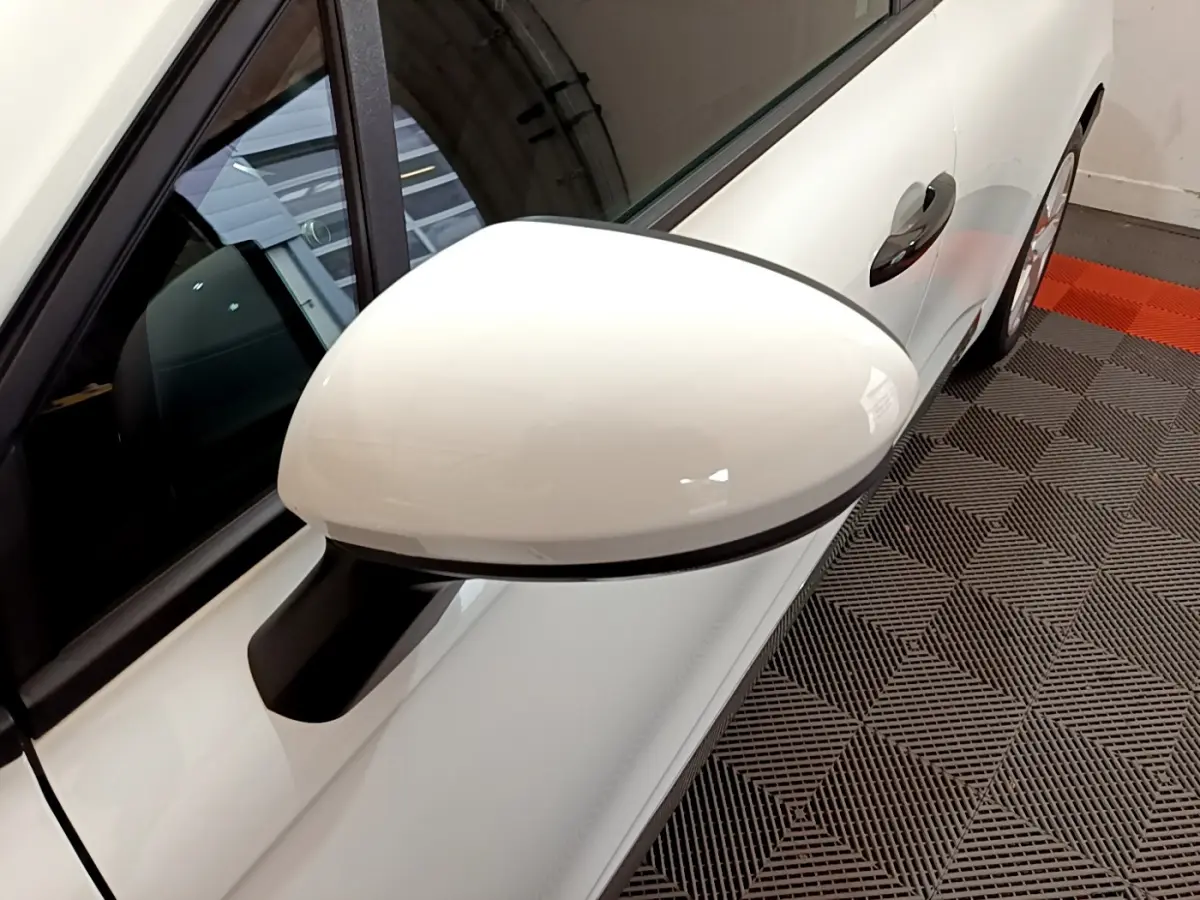 Rétroviseur blanc côté gauche de la Renault Clio Business 2021, avec poignée de porte visible sur fond de sol texturé.