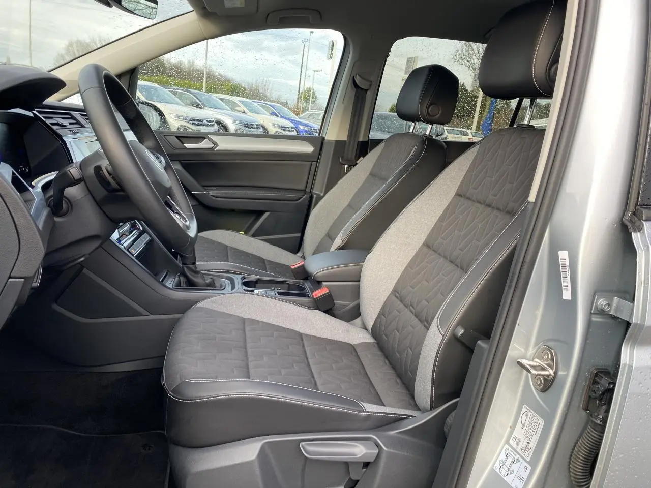 Vue intérieure côté conducteur du Volkswagen Touran 2025 en Oyster Argent métallisé, sièges tissu bicolore gris et noir.