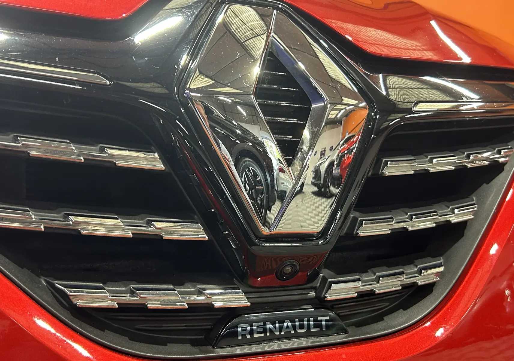 Gros plan sur la calandre rouge d'une Renault Clio V TCe 90 TECHNO avec logo chromé et caméra 360° intégrée.