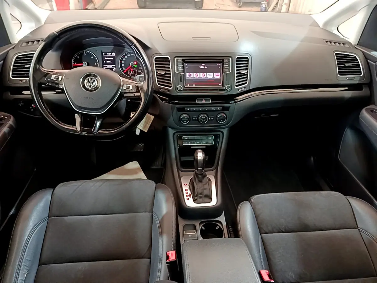 Vue intérieure avant du Volkswagen Sharan 2017 avec tableau de bord noir, volant multifonctions et console centrale avec boîte auto.