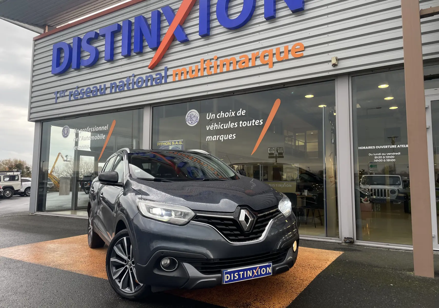 Renault Kadjar gris titanium en 3/4 avant droit devant un showroom Distinxion avec phares allumés et jantes alliage biton.