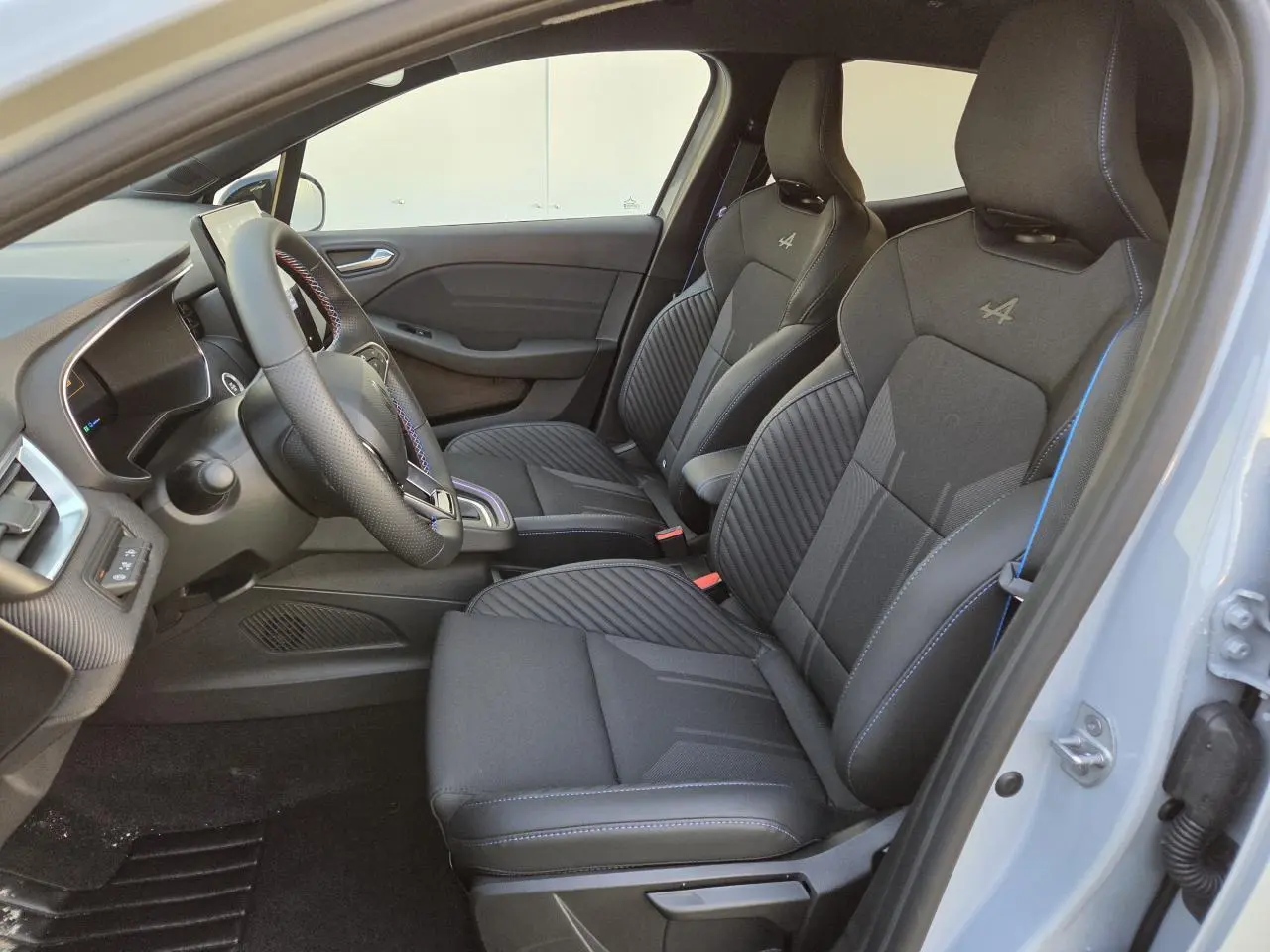 Intérieur avant de la Renault Clio V hybride, sièges sport noirs avec surpiqûres bleues et logo Alpine visibles côté droit.