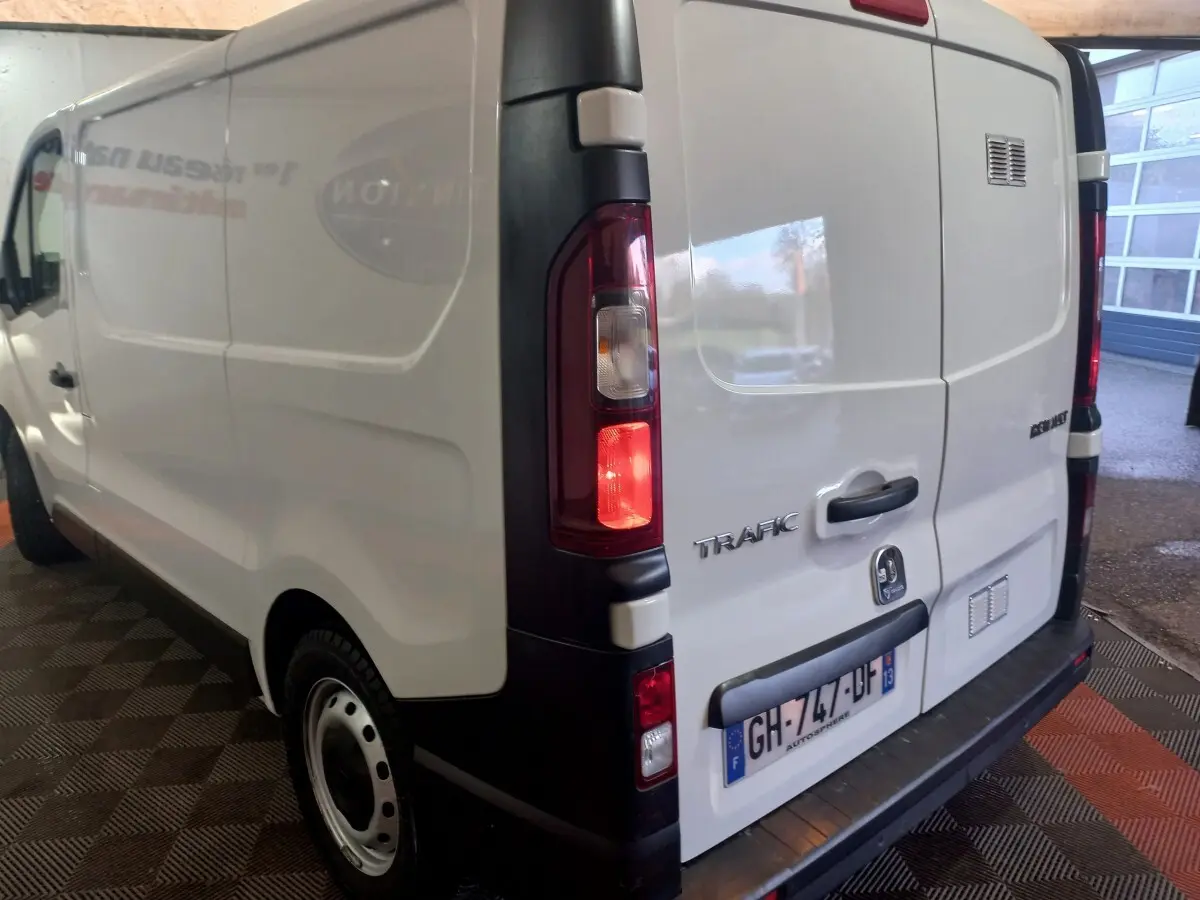 Vue 3/4 arrière gauche d'un Renault Trafic Fourgon blanc avec feux arrière allumés et plaque d'immatriculation française.