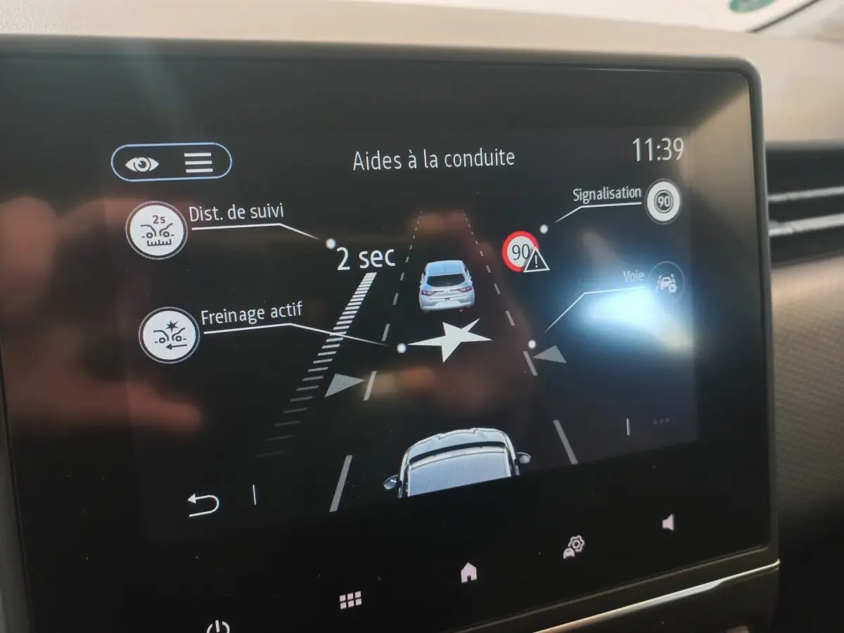 Écran tactile intérieur montrant l’aide à la conduite avec affichage de distance de suivi et freinage actif sur Renault Clio blanche.