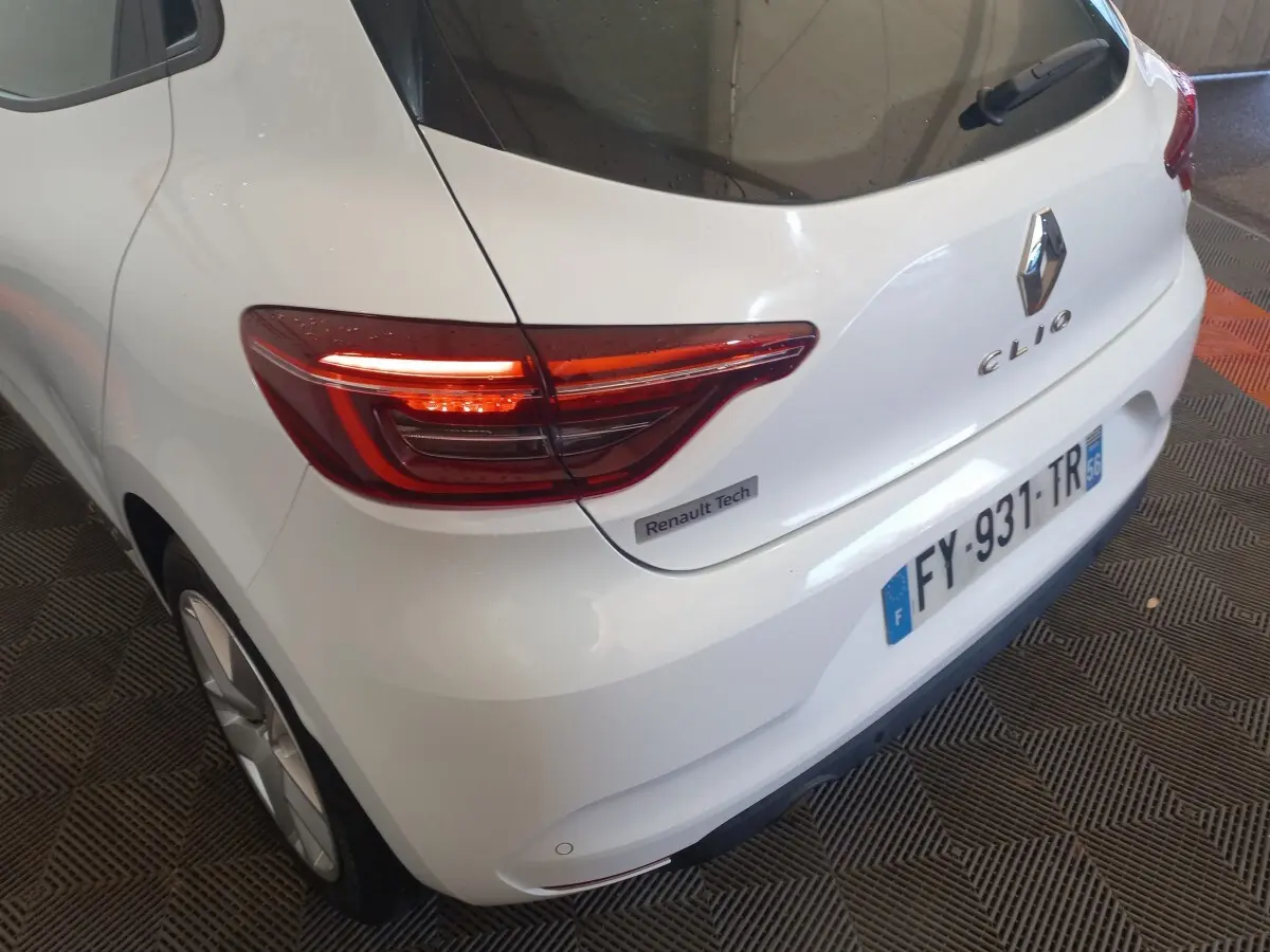 Vue 3/4 arrière droite d'une Renault Clio blanche avec feux arrière allumés et badge Renault Tech visible.