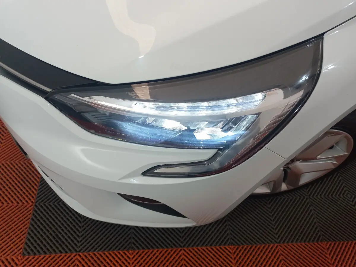 Gros plan sur le phare avant droit allumé d’une Renault Clio Business blanche, avec partie de la roue visible.