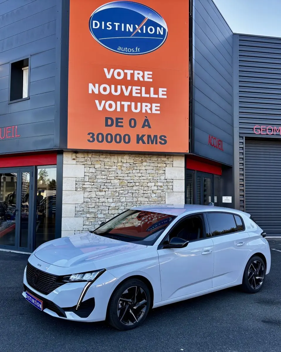 PEUGEOT 308 Hybrid blanc vue 3/4 avant droit, calandre noire et feux LED verticaux, devant un bâtiment commercial.