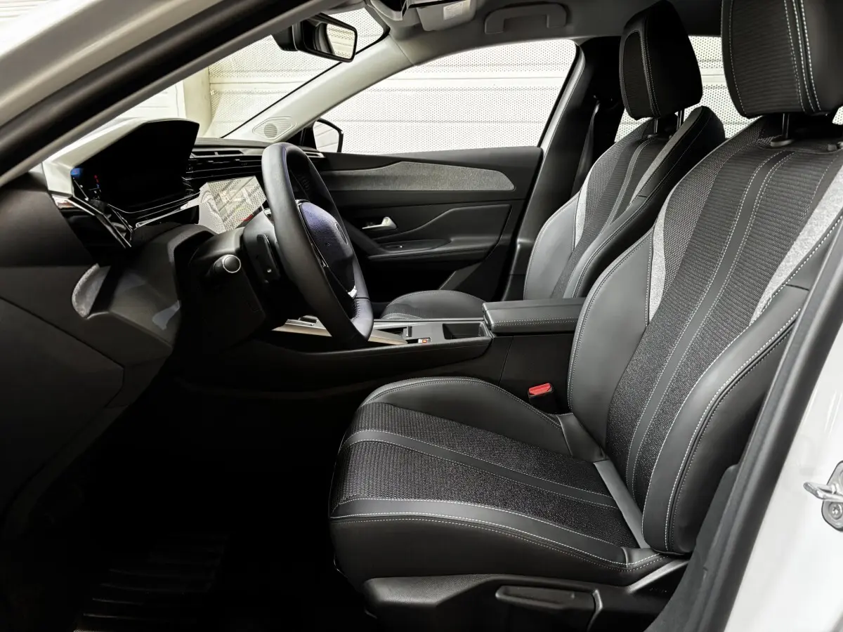 Vue intérieure côté conducteur de la Peugeot 308 Hybrid blanche, avec sièges noirs et volant cuir multifonctions.