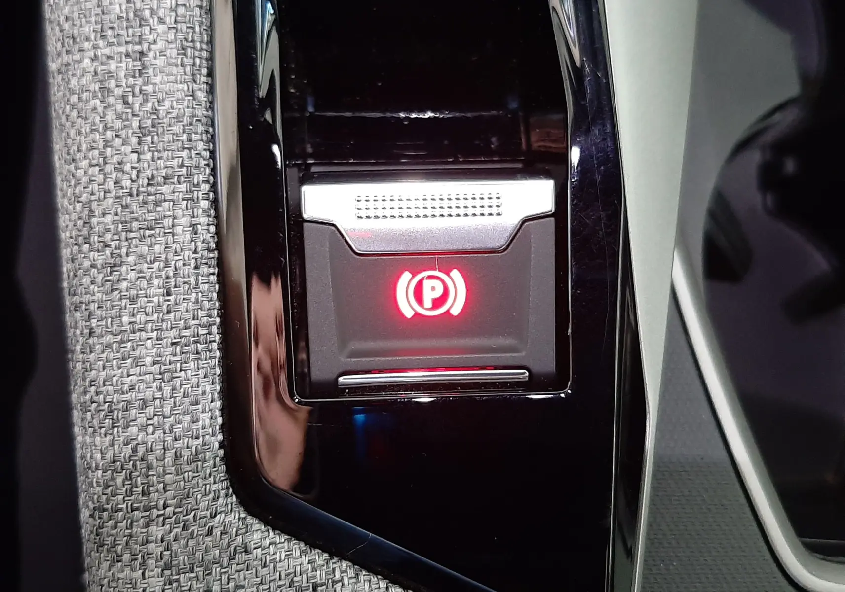 Gros plan sur le bouton de frein à main électronique rouge, entouré de garniture noire et tissu gris dans le Peugeot 3008 Bleu Ingaro.