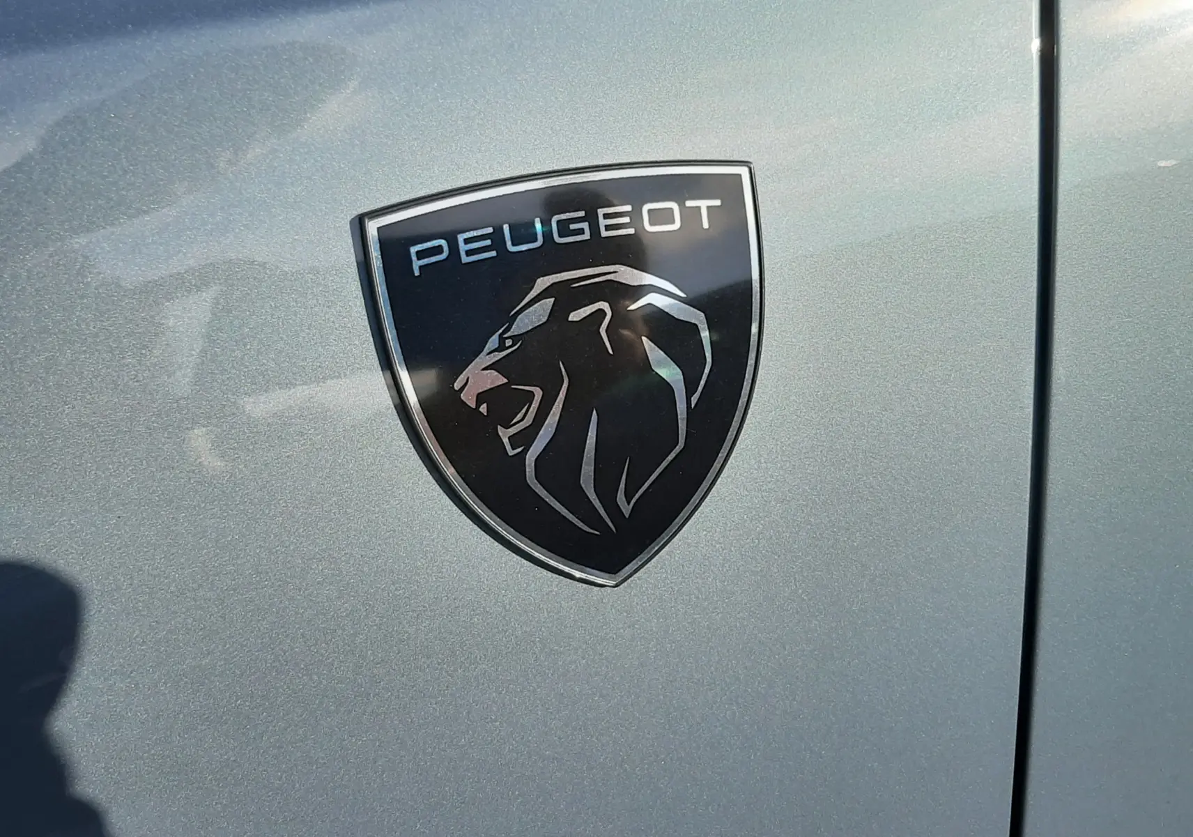 Gros plan sur l'écusson Peugeot noir et argent sur la carrosserie bleu Ingaro d'un 3008 hybride 2025.