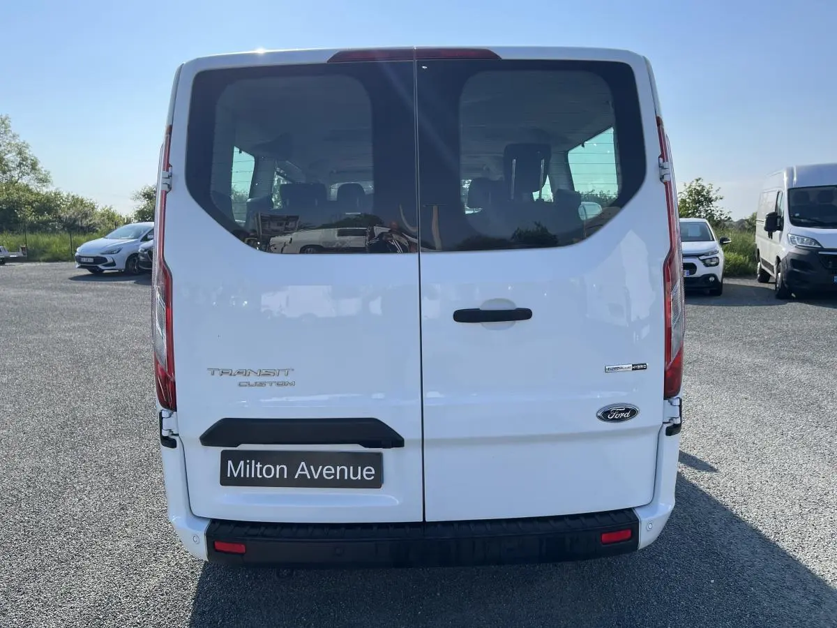 Vue arrière d'un Ford Transit Custom blanc avec hayon vitré et logo Ford visible sur un parking.