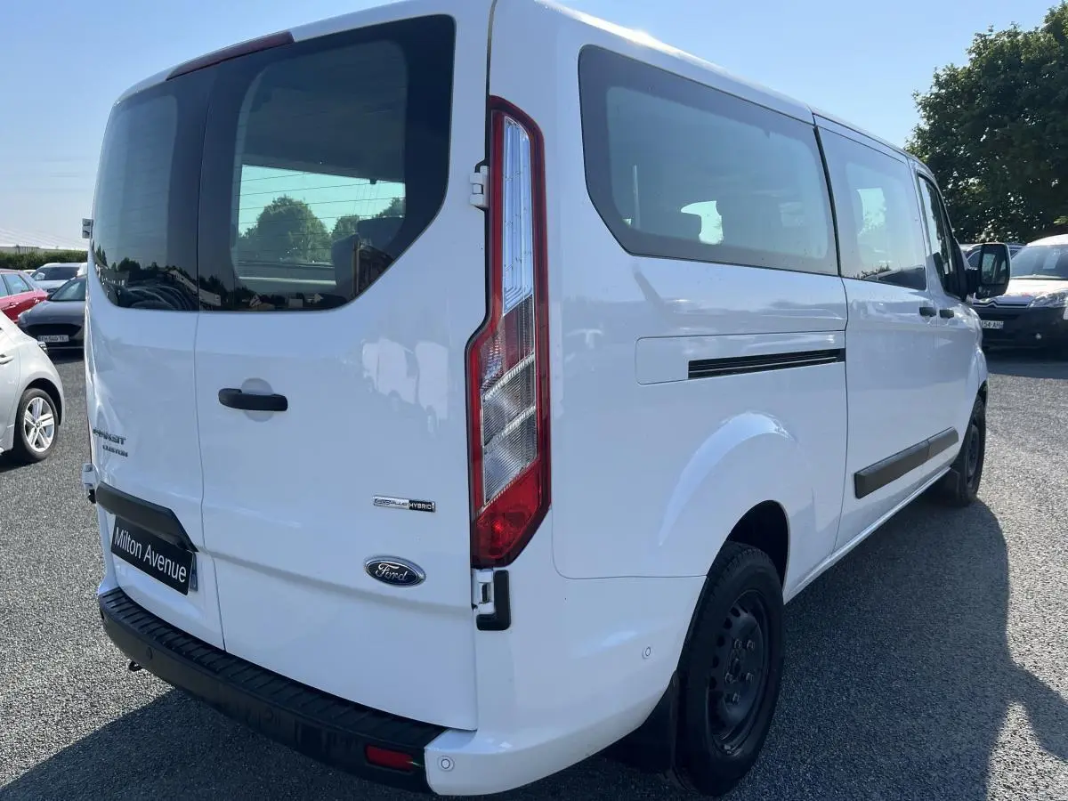 Vue 3/4 arrière droite d'un Ford Transit Custom Kombi blanc avec hayon vitré et badges Hybrid visibles.