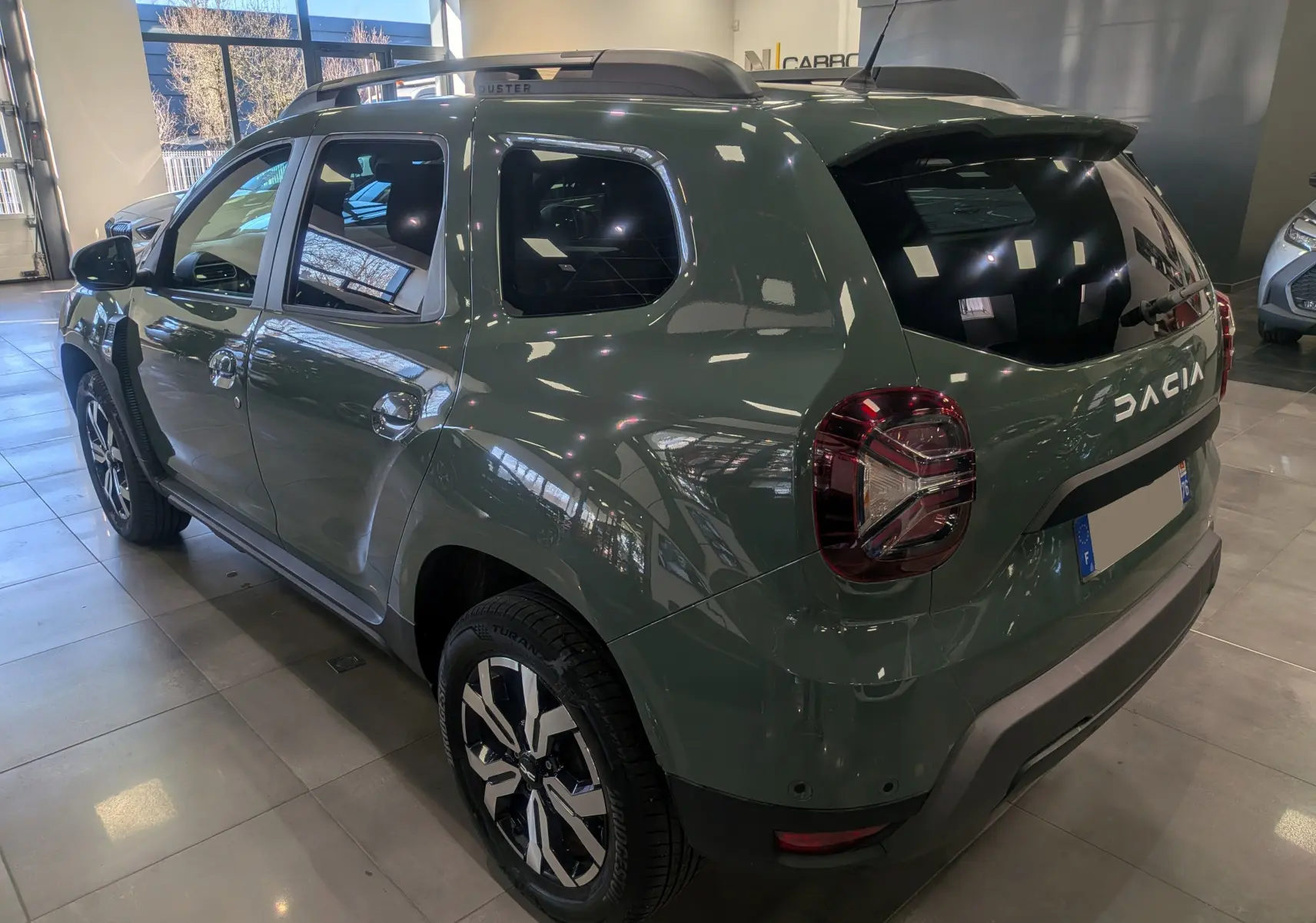 Vue 3/4 arrière droite du Dacia Duster 2023 kaki lichen avec jantes biton et barres de toit en showroom.