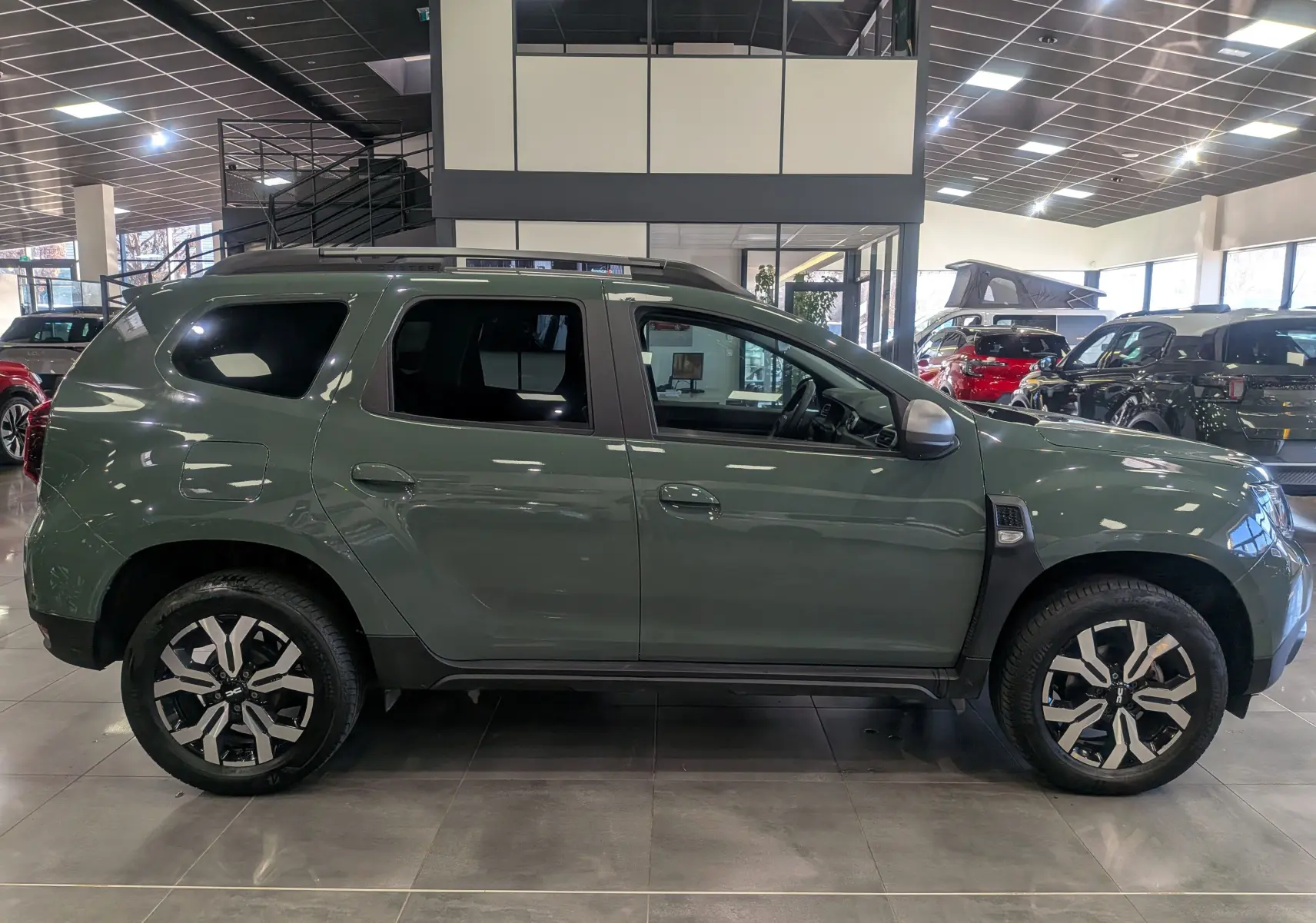 Vue de profil droit du Dacia Duster 2023 en kaki lichen, avec jantes alliage bicolores et barres de toit noires.