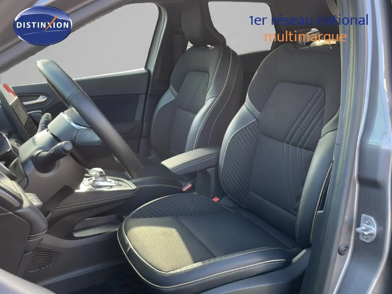 Vue intérieure côté conducteur de la Renault Symbioz gris cassiopée, sièges noirs avec surpiqûres blanches et volant visible.